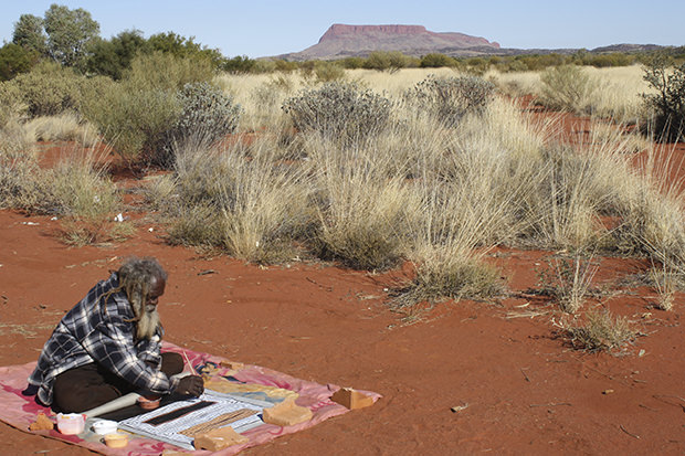 Artist Ronnie Tjampitjinpa painting on Country with the Kintore ranges behind him